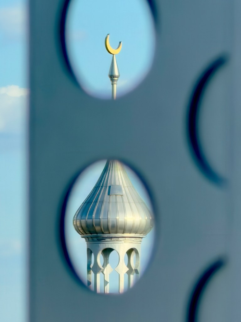 An architectural photograph by Amy Martz showing A minaret at UTampa in Tampa, Florida. Captured framing through two oval cutouts in a light blue foreground structure, the top oval highlights the gold crescent finial atop a slender silver spire, while the lower oval reveals the metallic silver onion dome and arched arcade of one of Plant Hall's iconic minarets against a clear blue sky under bright daylight.