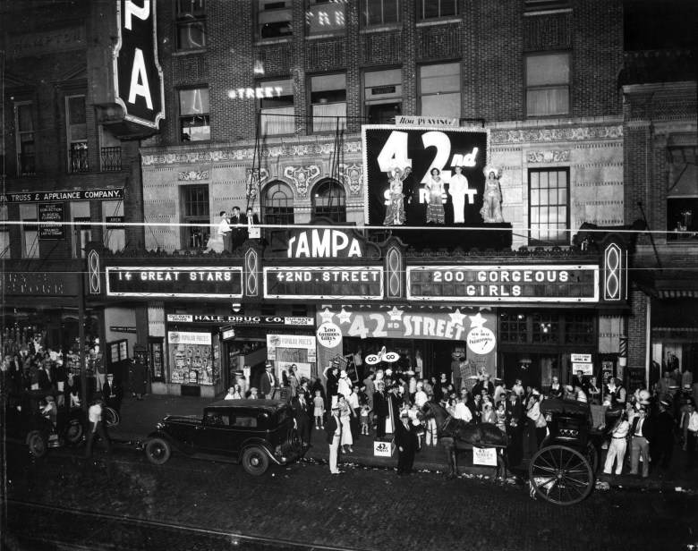 A historic black-and-white promotional photograph showing an exterior view of the Tampa Theatre in Tampa, Florida c. 1943, where a crowd is gathered for the opening night of '42nd Street.' Courtesy of USF Digital Commons, the wide marquee display reads "14 GREAT STARS", "42ND. STREET", and "200 GORGEOUS GIRLS" above a large illuminated rooftop sign featuring "42nd STREET" with costumed cutouts of performers. Below on the street, a large crowd in 1940s attire gathers around vintage automobiles and a horse-drawn carriage parked alongside the sidewalk in front of Hale Drug Co. and adjacent storefronts.