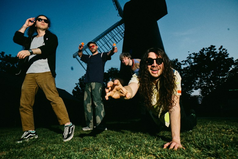 A low-angle outdoor promotional photograph by Joshua Tarn showing chiptune rock band Anamanaguchi posing on a grassy lawn with a large windmill in the background. In the foreground, a band member with long dark hair and sunglasses crawls toward the camera with an outstretched hand. Behind him, three other band members stand and pose in casual wear—including a white jacket, dark t-shirt, sunglasses, and sneakers—under a clear blue sky.