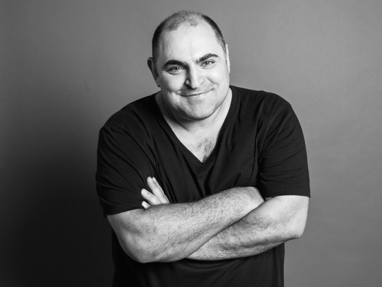 A black-and-white studio portrait photograph showing Australian electronic music artist and DJ Anthony Pappa posing against a solid grey background. Captured in a medium close-up, Pappa stands with his arms crossed over his chest, wearing a dark v-neck t-shirt and smiling directly into the camera under soft studio lighting.