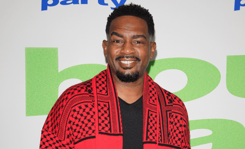 A medium portrait photograph by Joe Seer showing comedian and actor Bill Bellamy at the TCL Chinese 6 Theatres in Los Angeles, California on Jan. 11, 2023. Looking directly at the camera with a warm smile, Bellamy wears a black V-neck shirt underneath a red black-patterned open cardigan scarf or shawl. In the background, a white event step-and-repeat banner features green and blue lettering ("party").