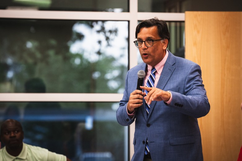 An indoor medium shot photograph by Dave Decker showing Tampa City Council member Luis Viera speaking at the C. Blythe Andrews Jr. Public Library in Tampa, Florida on July 9, 2026. Standing in front of indoor window panels and a light wood wall, Viera wears dark-framed glasses, a blue blazer, a pink button-down shirt, and a blue-and-white diagonal striped tie while speaking into a microphone held in his right hand and pointing forward with his left index finger. In the soft-focus background on the left, an audience member in a light green shirt observes the presentation under bright indoor lighting.