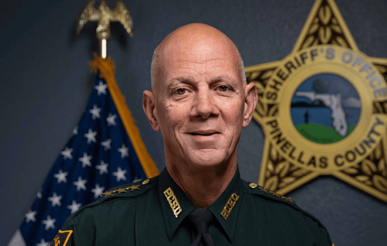 A close-up official portrait photograph of Pinellas County Sheriff Bob Gualtieri. Looking directly forward with a subtle smile, Gualtieri is shaved bald and wears a dark green Pinellas County Sheriff's Office uniform shirt with gold star insignia on the collar, gold shoulder embroidery, and a dark tie. Behind him on the left stands an American flag on a wooden flagpole with a gold eagle topper, and to his right, a large gold Pinellas County Sheriff's Office star badge seal is mounted on a grey wall.