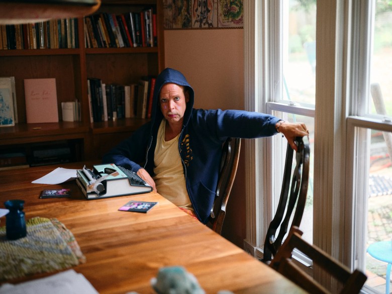 An indoor portrait photograph by Dave Kasnic showing singer-songwriter Bonnie 'Prince' Billy. Seated at a large wooden dining table in a brightly lit room next to double windows, he wears a light-colored undershirt beneath an unzipped navy blue hooded sweatshirt with the hood pulled over his head, resting his left arm across the back of a wooden chair. On the table before him sits an antique manual typewriter alongside papers and small objects, set against a background featuring a bookshelf filled with books and natural window light.