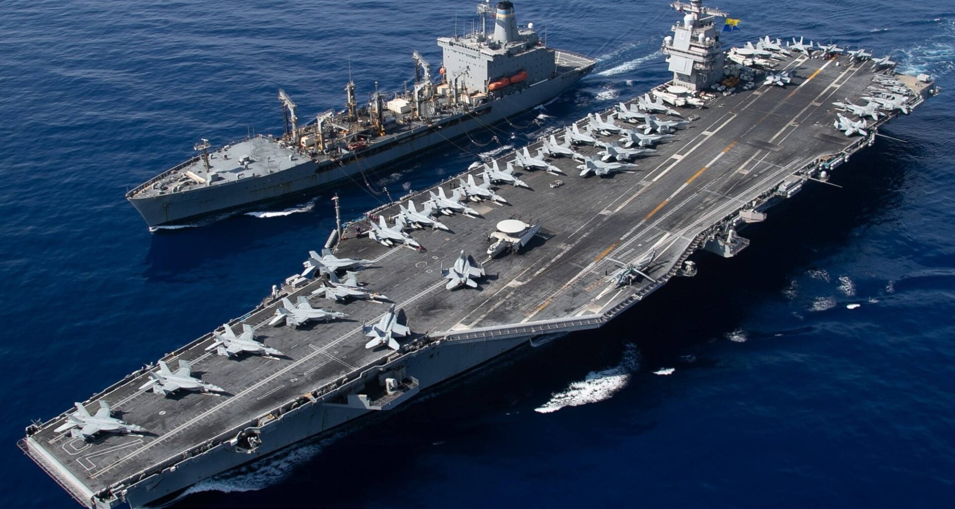 U.S. Navy aircraft carrier underway at sea alongside a replenishment ship, with aircraft positioned across the flight deck.