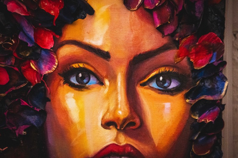 A close-up photograph by Dave Decker showing a colorful oil painting of a face by artist Carlos Pons inside Kress Contemporary in Ybor City, Florida on Sept. 17, 2026. Cropped across the nose, cheeks, and eyes, the portrait features warm golden skintones, deep blue expressive eyes, and vibrant red lips, surrounded by a crown of rich red, purple, and blue flower petals framing the top and sides of the canvas.