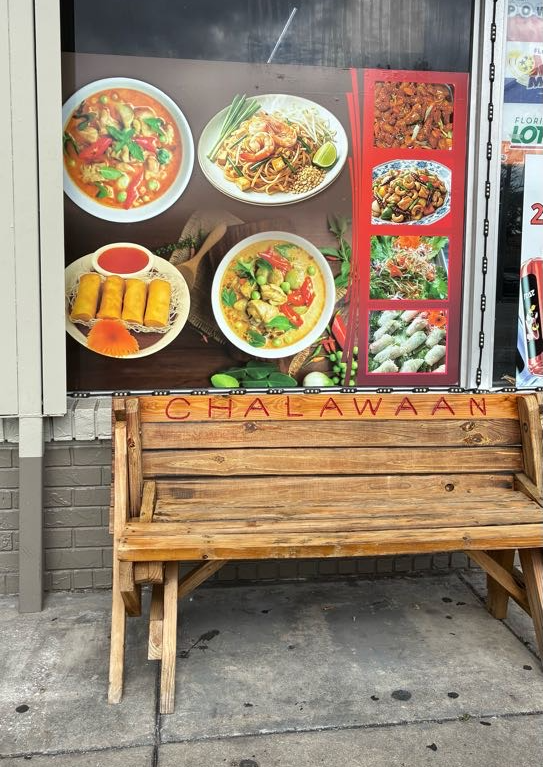 A high-angle exterior photograph by Annalise Mabe showing a wooden bench outside Chalawaan Thai Delish in Tampa, Florida on Aug. 21, 2026. In the center, a light-toned wooden picnic-style bench with "CHALAWAAN" carved across the top backboard sits on a concrete walkway against a grey brick wall. Directly behind the bench, a storefront window features colorful food decals depicting Thai dishes, including red and green curries, spring rolls, and Pad Thai.