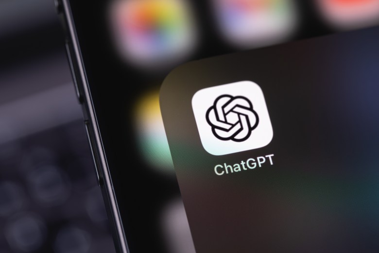 A extreme close-up shot of a smartphone screen displaying the ChatGPT app icon. The icon features a white stylized, interlocking circular logo on a black background, with the word "ChatGPT" printed in white underneath. The phone is held at an angle, showing blurred colorful app icons in the background and a portion of a laptop keyboard on the far left.