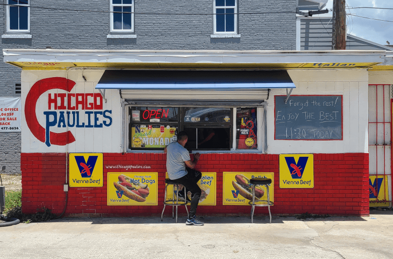 A wide exterior photograph by Ray Roa showing the Chicago Paulies food stand in Tampa, Florida on July 20, 2023. The small, single-story red and white building features a large painted "Chicago Paulies" logo on the left wall, Vienna Beef signs, yellow hot dog posters, and a chalkboard menu sign on the right. In the center, a person wearing a grey shirt sits on a stool at the ordering window beneath a dark retractable awning on a sunny day.
