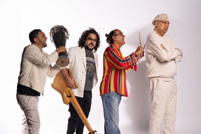 A promotional studio group photograph showing (L-R) Roberto Jr. Vizcaino, José Armando Golam Horacio "El Negro" Hernández, and Chucho Valdés. Standing against a plain white backdrop, four musicians are arranged in a playful line from left to right. On the far left, Roberto Jr. Vizcaino wears a cream jacket and grey pants while holding a djembe drum raised near his head. Next to him, José Armando Golam wears a white jacket over a graphic t-shirt and holds an acoustic bass guitar. Beside him, Horacio "El Negro" Hernández wears a colorful red, yellow, and black striped shirt with jeans, holding percussion sticks and laughing in profile. On the far right, Chucho Valdés stands facing forward with arms crossed, wearing a beige cap, a matching cream sweatshirt, and cream trousers under bright studio lighting.