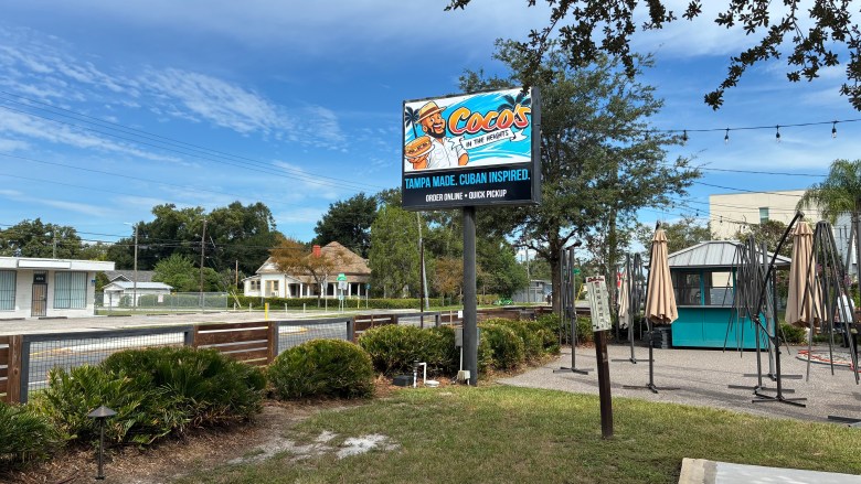 An outdoor landscape photograph by Annalise Mabe showing the roadside sign for Coco's In the Heights in Tampa, Florida on Sept. 29, 2026. Set against a bright blue sky with light wispy clouds, a tall black pole holds a prominent rectangular sign reading "Coco's" in colorful tropical lettering above text that reads "TAMPA MADE. CUBAN INSPIRED." Beneath the sign, a landscaped bed with green bushes and small lights sits along a wooden fence on the left. On the right, an outdoor patio area features closed patio umbrellas, string lights, and a small blue-and-white service hut set against surrounding trees.