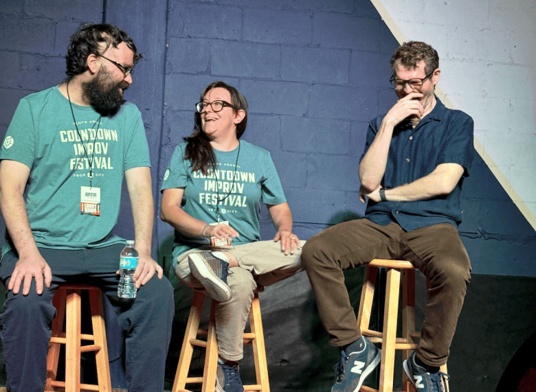 A medium group photograph showing (L-R) Justin Peters, Kelly Buttermore and Eddie Geller sitting on wooden barstools on stage at The Commodore in Ybor City, Florida on Aug. 7, 2026. On the left, Justin Peters sits holding a water bottle while wearing a beard, eyeglasses, dark trousers, and a teal "COUNTDOWN IMPROV FESTIVAL" t-shirt, smiling toward Kelly Buttermore in the center, who wears matching festival t-shirt, eyeglasses, khaki pants, and dark sneakers. On the right, Eddie Geller wears a dark button-up shirt, brown trousers, eyeglasses, and grey New Balance sneakers while resting his hand over his mouth and laughing, set against a painted brick backdrop.