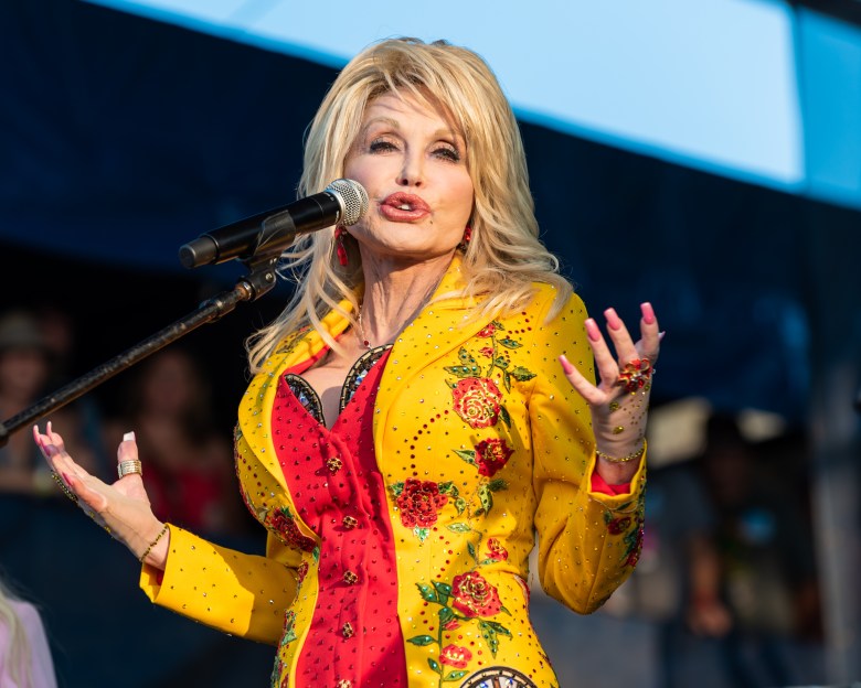 A stage performance photograph by Carl Beust showing Dolly Parton plays Newport Folk Festival at Fort Adams State Park in Newport, Rhode Island on July 27, 2019. Captured in a medium shot from a low angle under bright daylight, Parton stands on stage with styled, volumetric blonde hair, wearing a vibrant yellow long-sleeved jacket heavily embellished with red floral rose embroidery and rhinestones over a bright red corseted bodice. She sings into a dark microphone mounted on a stand in front of her while gesturing expressive, open-handed poses with both hands adorned with rings and red manicured nails.