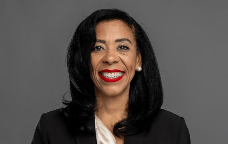 A close-up official portrait photograph of Dr. Patricia Alonzo. Looking directly forward with a wide, smiling expression and bright red lipstick, Alonzo has shoulder-length dark hair curled at the ends and wears a pearl earring. She is dressed in a dark blazer over a white collared blouse, positioned against a solid, neutral grey studio backdrop.