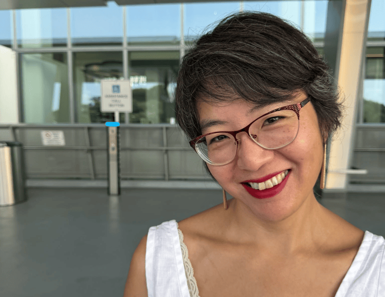 A close-up outdoor candid photograph of pianist Eunmi Ko. Smiling brightly toward the camera with red lipstick, Ko has short grey-streaked dark hair and wears tortoise-shell glasses, dangling drop earrings, and a white V-neck top with lace trim around the neckline. She is positioned on an outdoor covered walkway with large glass windows and metal architectural pillars in the background under natural daylight.