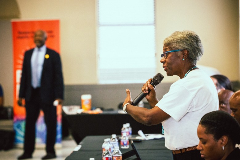 An indoor medium shot photograph by Dave Decker showing speaker Fran Tate speaking at the Florida Rising Forum at the Martin Luther King Recreation Center in Tampa, Florida on Aug. 16, 2025. Standing in profile at a long black table lined with bottled water, Tate wears blue-framed glasses, a white short-sleeved t-shirt, and a necklace while holding a microphone in her right hand and gesturing with her left. In the soft-focus background on the left, a man in a dark suit stands near a orange-and-blue "Florida Rising" banner, while audience members sit along the table in the foreground under bright interior lighting.
