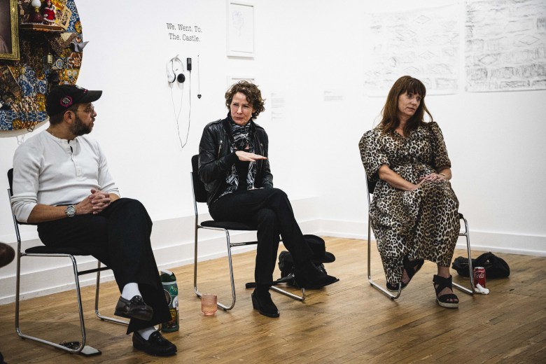 A wide indoor gallery photograph by Dave Decker showing (L-R) Danny Olda, Noelle Mason, and Liz Dimmitt at Tempus Projects in Ybor City, Florida on Sept. 17, 2026. Three panel speakers sit in a row on metal-framed chairs on a light wood floor against white art gallery walls. On the left, Danny Olda sits wearing a baseball cap, a long-sleeved light grey shirt, black trousers, and loafers while gesturing mid-conversation. In the middle, Noelle Mason sits wearing a dark leather jacket over a black patterned top, black trousers, and dark shoes, actively speaking with hand gestures. On the right, Liz Dimmitt sits in a leopard-print midi dress and dark shoes, resting her hands in her lap. Framed artwork, wall text, and headphones are displayed on the wall in the background.