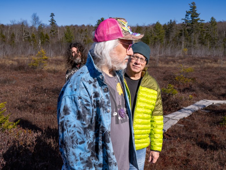 An outdoor band portrait photograph by Jeff Fowler showing (Front-back) J Mascis, Murph, and Lou Barlow of Dinosaur Jr.. Standing along a wooden boardwalk in an open bog landscape under bright daylight, frontman J Mascis stands prominently in the foreground profile facing right, wearing a bright pink mesh-backed baseball cap, glasses, a blue and black tie-dye jacket over a dark grey t-shirt, and a necklace. Behind him, bassist Lou Barlow stands facing forward wearing a dark beanie, glasses, and a neon lime-green quilted puffer jacket over a black shirt. In the background peeking behind Mascis, drummer Murph stands looking forward with long dark curly hair under a clear blue sky.