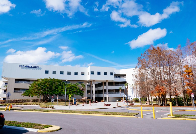 A wide exterior photograph showing the Technology Building at Hillsborough Community College's Dale Mabry campus on Jan. 25, 2021. Captured under a bright blue sky with light clouds, the white multi-story academic building features large windows and prominent dark lettering reading "TECHNOLOGY" on its upper left facade. In the foreground, an asphalt driveway and parking area lead past a green lawn, leafy trees, and reddish cypress trees on the right side of the campus grounds.