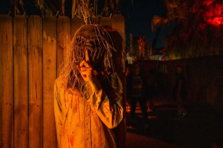 A dramatic, low-light night photograph of a scare actor performing during Howl-O-Scream at Busch Gardens Tampa Bay in Tampa, Florida. Lit by warm orange spotlighting against a dark wooden fence, a costumed figure stands centered while wearing a long-sleeved, blood-stained shirt and a tattered burlap mask with hanging frayed strands covering their face. The figure holds an index finger up to their lips in a "shh" hushing gesture, set against dark shadowy backgrounds and red accent lighting in the distance.