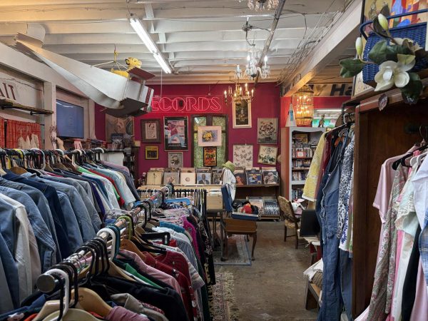The room shows clothes on racks, paintings on a magenta wall, and chairs in the iddle of the room