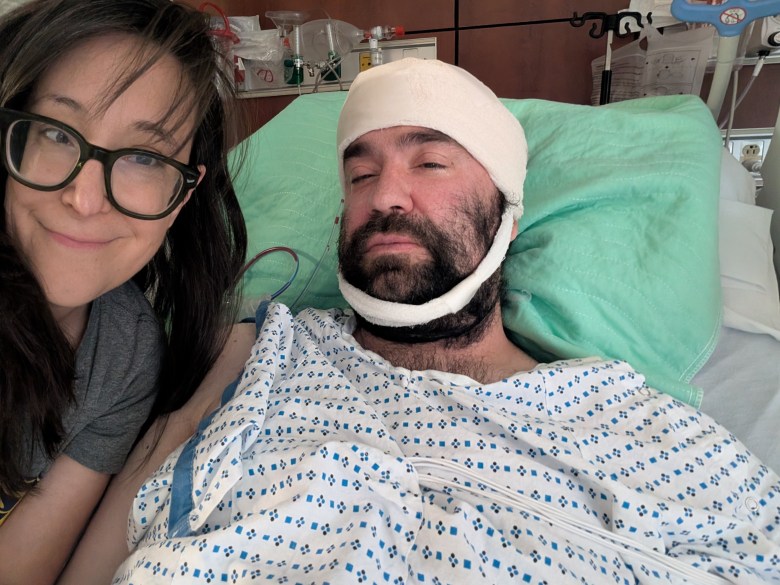 A close-up selfie photograph showing comedian Kelly Buttermore sitting alongside Justin Peters in a hospital room at Lenox Hill Hospital in New York City, New York on June 8, 2026. On the left, Buttermore smiles gently toward the camera while wearing thick black-rimmed eyeglasses and a grey t-shirt. On the right, Peters lies in a hospital bed wearing a patterned blue and white patient gown, resting against a light green pillow with a thick white medical bandage wrapped around his head and chin. Medical monitoring equipment, wires, and wall-mounted cabinets are visible in the background behind the bed.