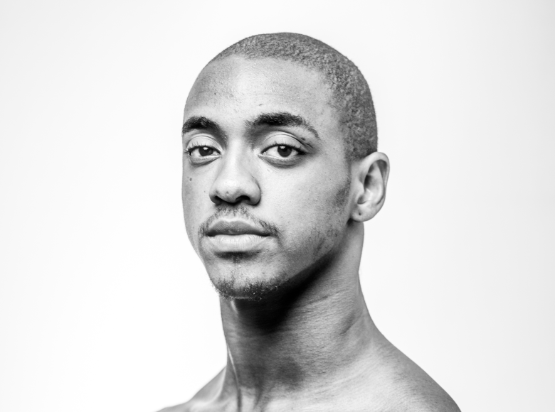 A black-and-white studio headshot portrait showing dancer Jhon Destrade. Captured in a close-up three-quarter angle against a seamless light grey background, Destrade appears bare-shouldered while looking directly into the camera with a neutral, poised expression. He has short closely cropped hair, dark eyes, and subtle facial hair along his upper lip and chin under soft, even studio lighting.