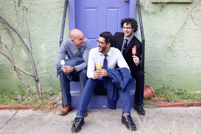 An outdoor promotional portrait photograph showing jazz trio (L-R) John O'Leary III, Alejandro Arenas, and Mark Feinman of La Lucha. Seated together on the concrete steps of a purple exterior doorway against a pale green building wall, the three musicians dress in suits while holding ice cream popsicles and laughing together. On the left, John O'Leary III wears a grey suit jacket, blue trousers, and brown shoes while holding a white popsicle. In the center, seated a step lower, Alejandro Arenas wears a white dress shirt, dark tie, blue trousers, and black dress shoes while holding a yellow popsicle and resting his suit jacket on his knee. On the right, Mark Feinman sits wearing a dark black suit, dark tie, and black shoes while holding a red popsicle under bright daylight.