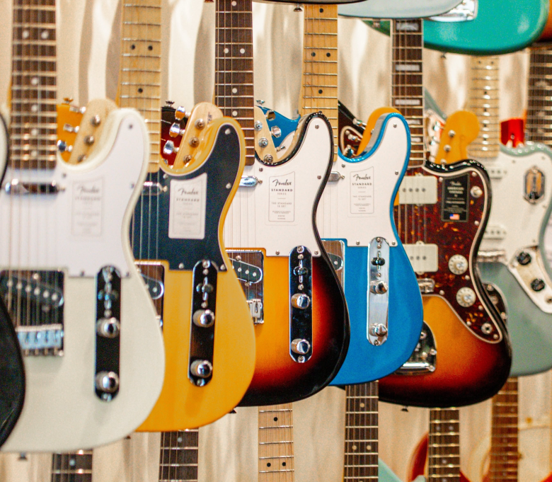 A close-up photograph showcasing a row of Fender electric guitars hanging on display at Legends Guitars in Tampa, Florida. Captured at an angled perspective along the store rack, the foreground features a white Telecaster, a butterscotch blonde Telecaster, a three-tone sunburst Telecaster, a bright blue Telecaster, and a sunburst Jaguar with a tortoiseshell pickguard, alongside additional electric guitars suspended in the background against a light wooden wall.