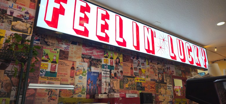 An interior photograph by Ray Roa showing the ordering counter and sign inside Lucky Tigre in Tampa, Florida on Aug. 9, 2025. Mounted above the counter, a illuminated rectangular light box sign reads "FEELIN' LUCKY?" in bold red, retro-style block lettering with stylized starbursts. Below the sign, a vibrant wall is completely covered in a collage of vintage posters, ads, and news clippings, flanked by hanging greenery on the left.