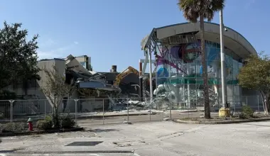 A crane is shown tearing down a mall building surrounded by a chain-link fence.