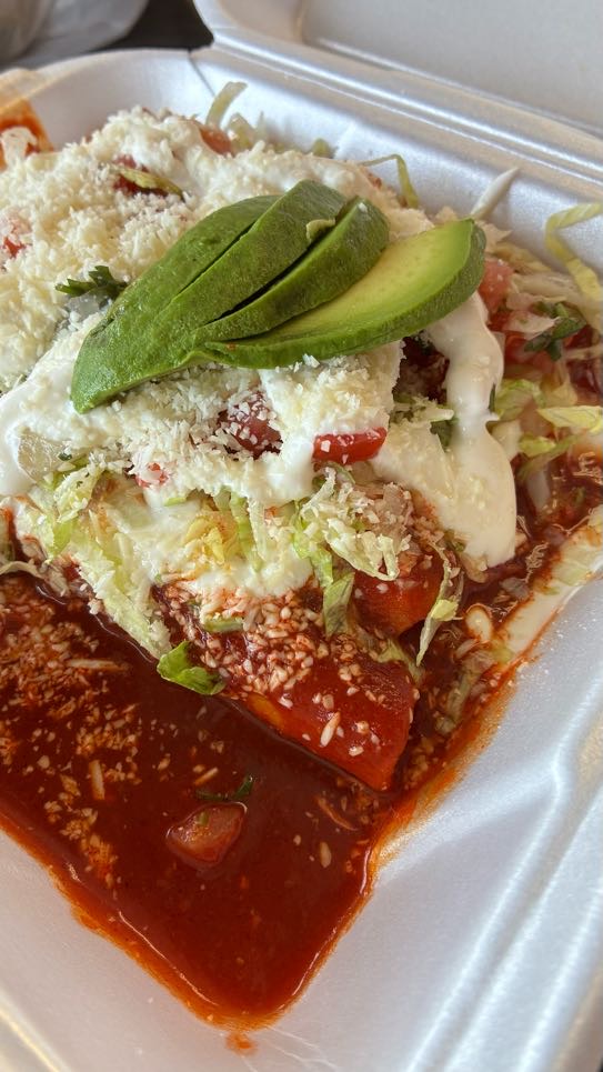 A close-up food photograph by Annalise Mabe showing a serving of enchiladas at Mangonadas in Tampa, Florida on Aug. 22, 2026. Served inside a white foam takeout container, rolled enchiladas are smothered in a rich, pool-like red chili sauce and topped with shredded lettuce, diced tomatoes, a heavy layer of crumbled white cotija cheese, drizzled white crema, and fan-sliced fresh green avocado.