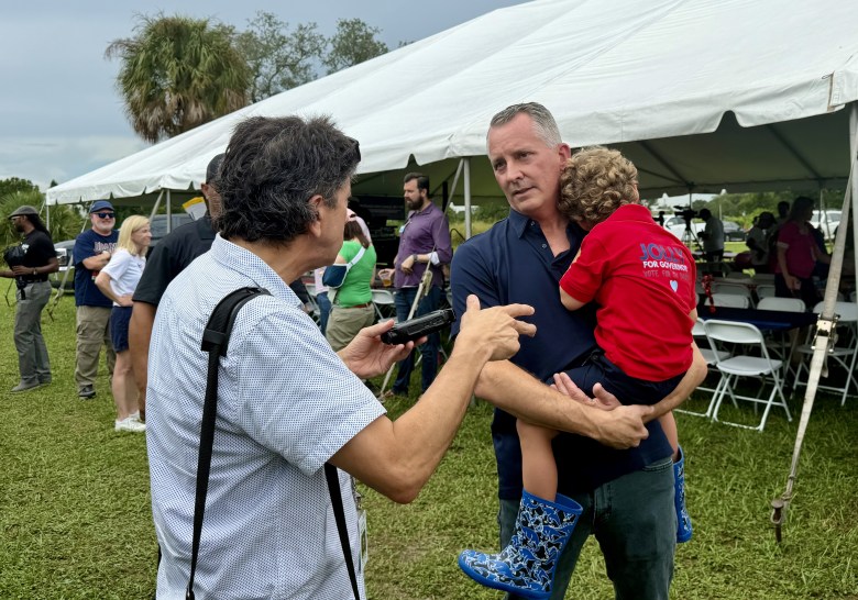 A candid outdoor photograph showing journalist Mitch Perry interviewing David Jolly at IBEW Local 915 in Tampa, Florida on Sept. 7, 2026. On the left, Perry stands in profile wearing a short-sleeved grey button-up shirt and a black shoulder strap, holding a small recording device and gesturing toward Jolly while speaking. On the right, Jolly wears a dark navy polo shirt while holding a young child wearing a red shirt and blue patterned rain boots, who rests their head on his shoulder. In the background, an outdoor gathering takes place under a large white event tent on a grass field under an overcast sky.