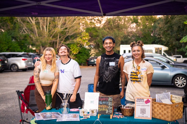 A wide exterior photograph showing vendors standing behind their booth at the New Moon Makers Market in St. Petersburg, Florida. Under a purple pop-up canopy, four smiling organizers and makers stand behind a table covered in promotional flyers, small signs, a woven basket, and a chalkboard sign. On the far left, a person with blonde hair wears a crop top and brown pants next to a person in a white t-shirt. In the center, a person wearing a black sleeveless top stands beside a person on the right wearing a cream-colored "New Moon Makers Market" t-shirt, set against an outdoor parking lot with parked cars and trees in soft evening light.