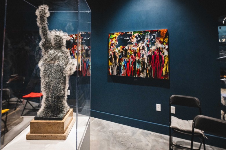 An indoor art gallery photograph showing artist Tatiana Mesa Paján's 'Libertad' at OXH Gallery in Tampa, Florida on Sept. 24, 2026. Displayed inside a clear glass acrylic pedestal case on the left, a silver wire mesh sculpture depicting the Statue of Liberty stands on a wooden base with raised arm and torch. Mounted on the dark blue gallery wall in the background is a colorful abstract painting filled with densely layered figurative and floral forms in red, green, yellow, black, and white. To the right, black folding chairs sit along the concrete floor beneath spotlighting.