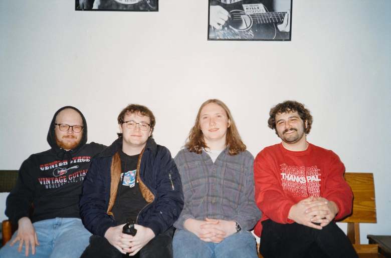 A promotional indoor flash photograph of the band Pat & The Pissers. Four band members sit side-by-side on a wooden bench against a plain white wall framed by small hanging picture frames above. From left to right: a man with a beard, glasses, and a black hood wearing a black graphic hoodie and blue jeans; a man wearing glasses, a dark jacket over a graphic t-shirt, and dark trousers; a band member with long straight light-brown hair wearing a grey flannel shirt and blue jeans with hands folded in their lap; and a man with short curly dark hair and a mustache wearing a red sweatshirt with white text across the chest.