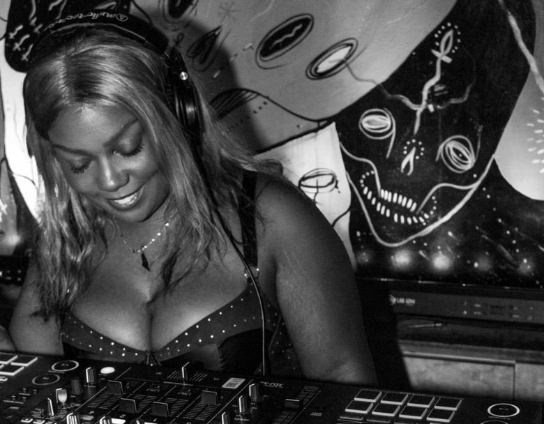 A black-and-white indoor portrait photograph by Jen Bodine showing DJ Post-Physique pictured at The Bends in St. Petersburg. Captured from a medium close-up shot while performing behind a DJ controller turntable console, Post-Physique smiles downward toward the equipment controls while wearing over-ear headphones, a delicate pendant necklace, and a dark studded bustier top. In the background behind the DJ booth, stylized dark artwork depicting a skull motif is illuminated under ambient venue lighting.