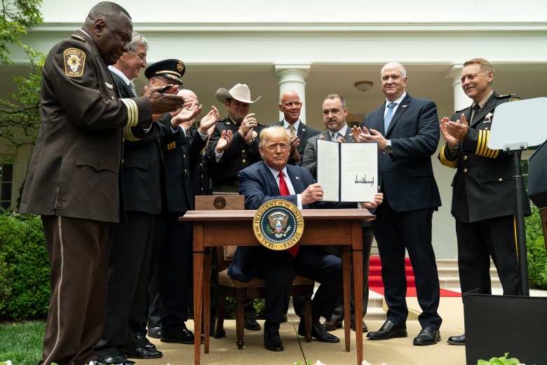 A medium shot photograph showing President Donald J. Trump, with Pinellas Sheriff Bob Gualtieri (center, rear) signing an executive order on safe policing for safe communities on June 16, 2020, in the Rose Garden of the White House in Washington, D.C. Seated at a wooden desk adorned with the presidential seal, President Trump holds up the signed document while surrounded by a group of law enforcement officials and suit-clad dignitaries clapping in applause, including Sheriff Bob Gualtieri standing in the rear center in a suit.