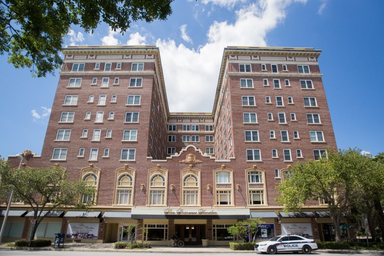 A wide exterior architectural photograph showing The Princess Martha building in St. Petersburg, Florida c. 2017. Captured from street level across the road, the U-shaped, multi-story historic red brick building features classical archways, decorative stone detailing along the facade, and large ground-floor storefront windows beneath a street-level awning. A white police cruiser is parked along the curb on the right, with green leafy trees framing both sides of the building under a clear blue sky with light white clouds.