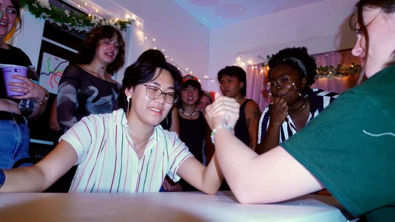 An indoor event photograph showing participants during A Queer Expressions arm wrestling fundraiser in St. Petersburg, Florida on June 21, 2024. Captured from a medium shot at table height under cool-toned party lighting, a person on the left wearing glasses and a white button-down shirt with vertical red and blue stripes smiles while arm wrestling an opponent on the right in a dark green t-shirt. A crowd of onlookers watches from behind the table, including a person in a black patterned long-sleeve top, a spectator in a black tank top and baseball cap, and a person on the right wearing glasses and a dark headband watching intently with a hand near their chin. Warm string lights and floral garlands hang along the wall in the background.