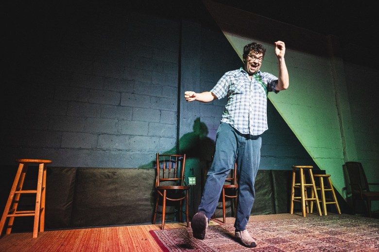 An indoor, medium-shot photograph of performer Justin Peters on a small stage at The Commodore in Tampa. Peters is caught in an energetic, mid-movement stance, leaning slightly to their left with one leg kicked out and arms outstretched. They have short, dark, tousled hair and are wearing a light-colored, short-sleeved plaid button-down shirt tucked into dark blue jeans. They are smiling broadly at the audience. The stage is sparsely furnished with two wooden stools and a small wooden chair. The background is a dark, painted cinder-block wall, and the stage floor is covered with a patterned, worn area rug. The scene is illuminated by warm, focused stage lighting.