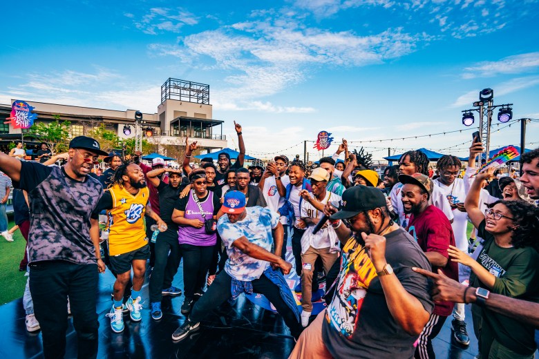 A high-energy, wide-angle photograph by Yvonne Gougelet of dancers and participants auditioning at the Red Bull Dance Your Style Qualifier at Armature Works in Tampa, Florida on April 9, 2023. Captured outdoors on a sunny day under a bright blue sky with light clouds, a crowd of street dancers cheer and react while surrounding performers in a dance cipher on a dark floor mat. On the left, a dancer in a dark patterned top and black baseball cap extends his arm outward in excitement. In the center foreground, a dancer wearing a blue graphic t-shirt and blue baseball cap drops low into a dynamic dance stance while surrounded by smiling spectators, performers in colorful graphic tees, and outdoor event lighting trusses.