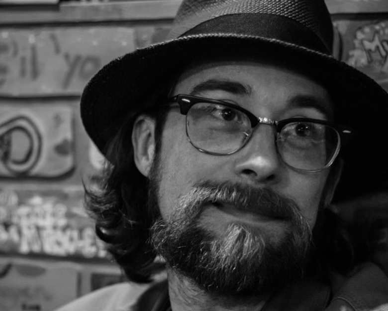 A black-and-white close-up portrait photograph of Richard J. Girard II. Looking slightly to his left with a gentle smile, Girard wears dark horn-rimmed eyeglasses, a dark fedora-style woven hat, a full beard and mustache, and shoulder-length wavy hair. In the background, a brick wall covered with faint graffiti and lettering is visible behind his right shoulder.