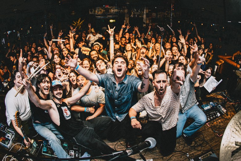 A high-energy live concert photograph showing indie rock band Rohna posing on stage with a cheering crowd in the background at Crowbar in Ybor City, Florida on May 15, 2026. Captured from a low front-of-stage angle, band members kneel and crouch together while raising their arms, surrounded by stage equipment, pedalboards, and drum cymbals as an enthusiastic audience behind them holds up hand signs and cheers.