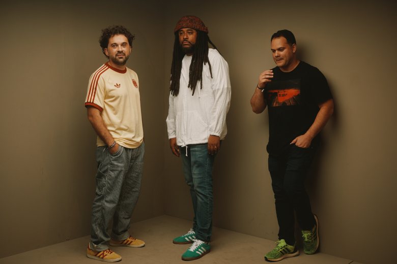 A promotional group portrait of (L-R) Rum & Coke DJs Harold Fandino, James Alvarez-Bacon (DJ Kumi), and Jesus Rodriguez posing against a muted grey corner wall backdrop. On the left, Harold Fandino wears a cream Adidas t-shirt with red sleeve stripes, blue jeans, and yellow sneakers. In the center, James Alvarez-Bacon (DJ Kumi) wears a maroon beanie, a white long-sleeved quarter-zip jacket, blue jeans, and green sneakers with white stripes while facing forward with dreadlocks. On the right, Jesus Rodriguez wears a black graphic t-shirt, dark pants, and green sneakers with yellow laces, looking slightly downward with one hand near his neck.