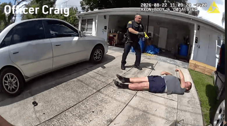A bodycam screenshot showing A still from bodycam footage showing St. Petersburg Police officer Daniel Poveda and Brian Pepal in St. Petersburg, Florida on Aug. 22, 2026. Captured in a wide-angle perspective on a concrete driveway outside a residential home, a uniformed police officer in dark attire stands with a yellow conductive energy device (Taser) pointed toward a man lying on his back on the pavement wearing a grey t-shirt and dark shorts. A white sedan is parked along the left side of the driveway, and timestamp text reading "2026-08-22 11:09:20" appears in the upper right corner of the frame under bright daylight.