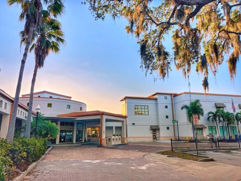 An outdoor landscape photograph by photographer Holly Apperson showing the exterior of the Safety Harbor Public Library in Safety Harbor, Florida. Captured under a clear blue sky illuminated by warm, late-afternoon sunlight, the multi-story stucco building features a drive-up covered entrance canopy on the left with palm trees and a Spanish-tile roof. Moss-draped oak branches frame the upper right foreground above a black metal fence, manicured green lawn, and parking lot in front of the building's main entrance.