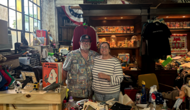Eric (left) has a white beard and wears a black baseball hat and a grey and white patterned shirt. Christina (right) is wearing a black and white striped shirt. Both are smiling.