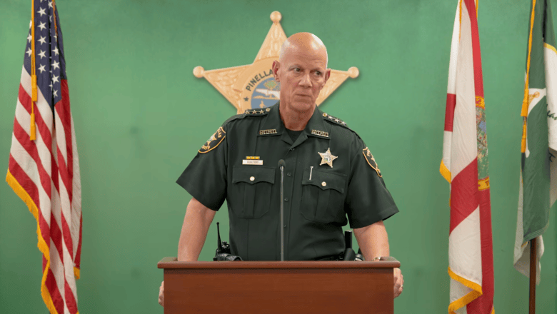 A screengrab from a Sept. 28, 2026 press conference led by Pinellas Sheriff Bob Gualtieri. Captured in a medium shot behind a dark wooden podium, Pinellas County Sheriff Bob Gualtieri stands wearing a dark law enforcement uniform with shoulder patches, a name badge, and a silver star emblem. He stands centered before a green backdrop featuring a large gold sheriff's star logo, flanked by the American flag on the left and the Florida state flag on the right under indoor press room lighting.