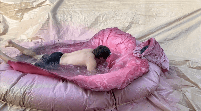 A high-angle production still showing a male performer lying in a pool of water in a short film from 'Squirter' which screened as part of the touring 2026 'Hump!' Film Festival. Lying face-down in a shallow pink-tarped pool filled with water, a man with light skin wearing black swim trunks rests his upper torso and chin near the edge of the enclosure. Wet ripples fold around his waist and legs against a textured, crushed-pink background under soft indoor lighting.